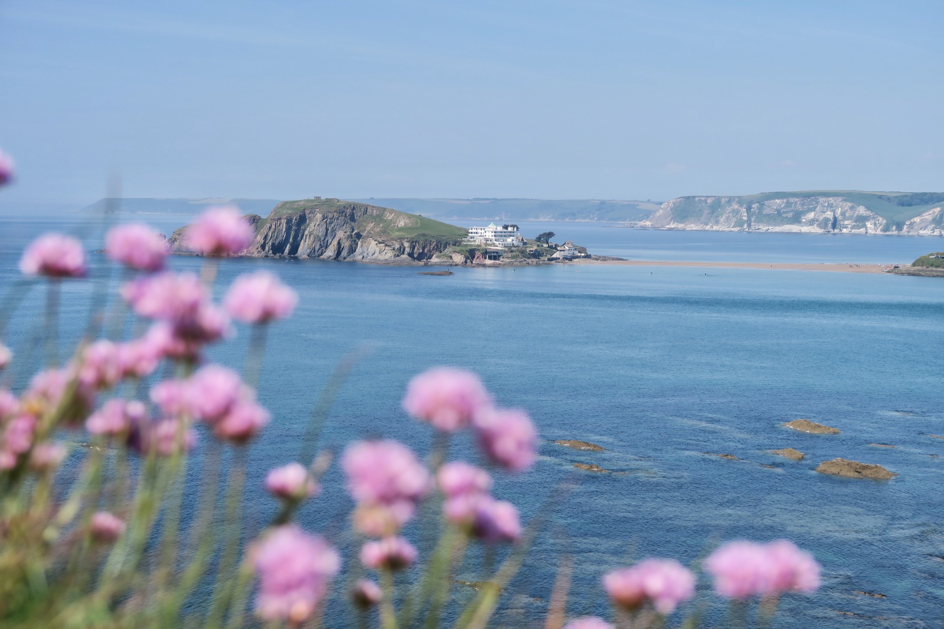 Burgh Island Devon. Island in the middle of a blue sea with a hotel on it. Pink flowers in foreground. 17 Beautiful Places to Visit in Devon for a Great Day Out. Devon England. Devon UK. Things to do in Devon. Places to see in Devon. What to see in Devon. Things to see in Devon. What to do in Devon. Devon attractions. Devon top attractions. Devon travel blog. Devon travel guide. The English Riviera. Exeter. Plymouth. Dartmouth. Dartmoor National Park. Exmoor National Park. Salcombe. Clovelly. Totnes. Appledore. Watermouth. Croyde. Woolacombe. Dartmouth. Ilfracombe. Beer. Burgh Island. Lundy Island. Click through to read more...