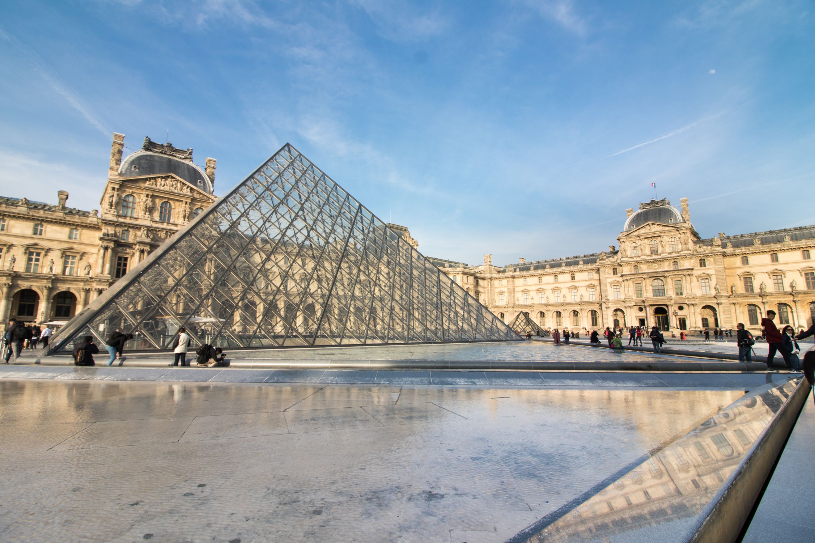 Louvre Museum and Art Gallery Paris. Glass pyramid in square with historic buildings and blue skies in background. Fountains in foreground.