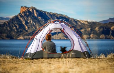 woman-in-tent-with-dog-camping-on-the-grass-near-a-lake-and-mountains-sustainable-travel-tips