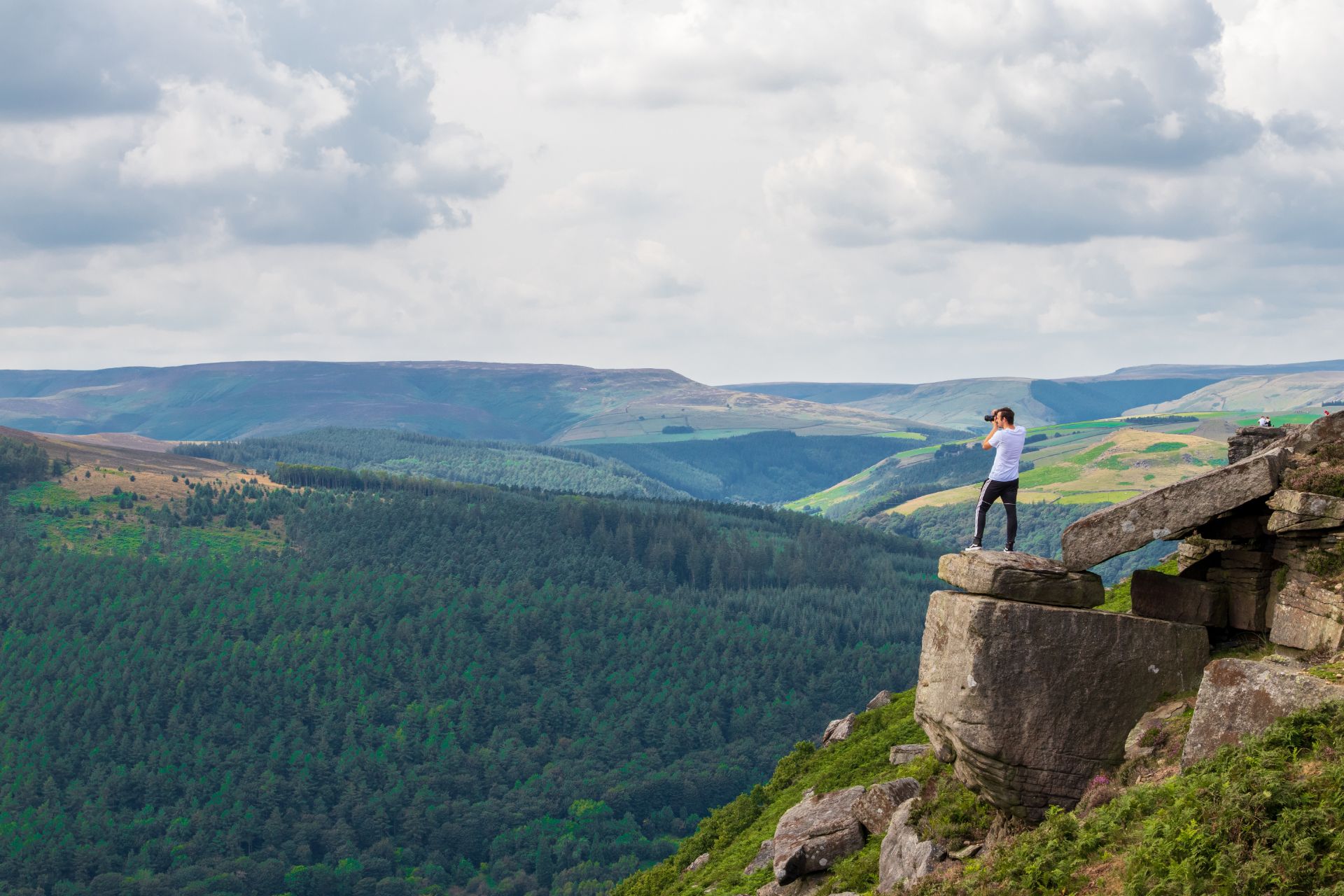man-standing-on-a-dangerous-rock-edge-on-a-hill-taking-a-photo-across-the-forests-to-his-left-at-bamford-edge-in-the-peak-district