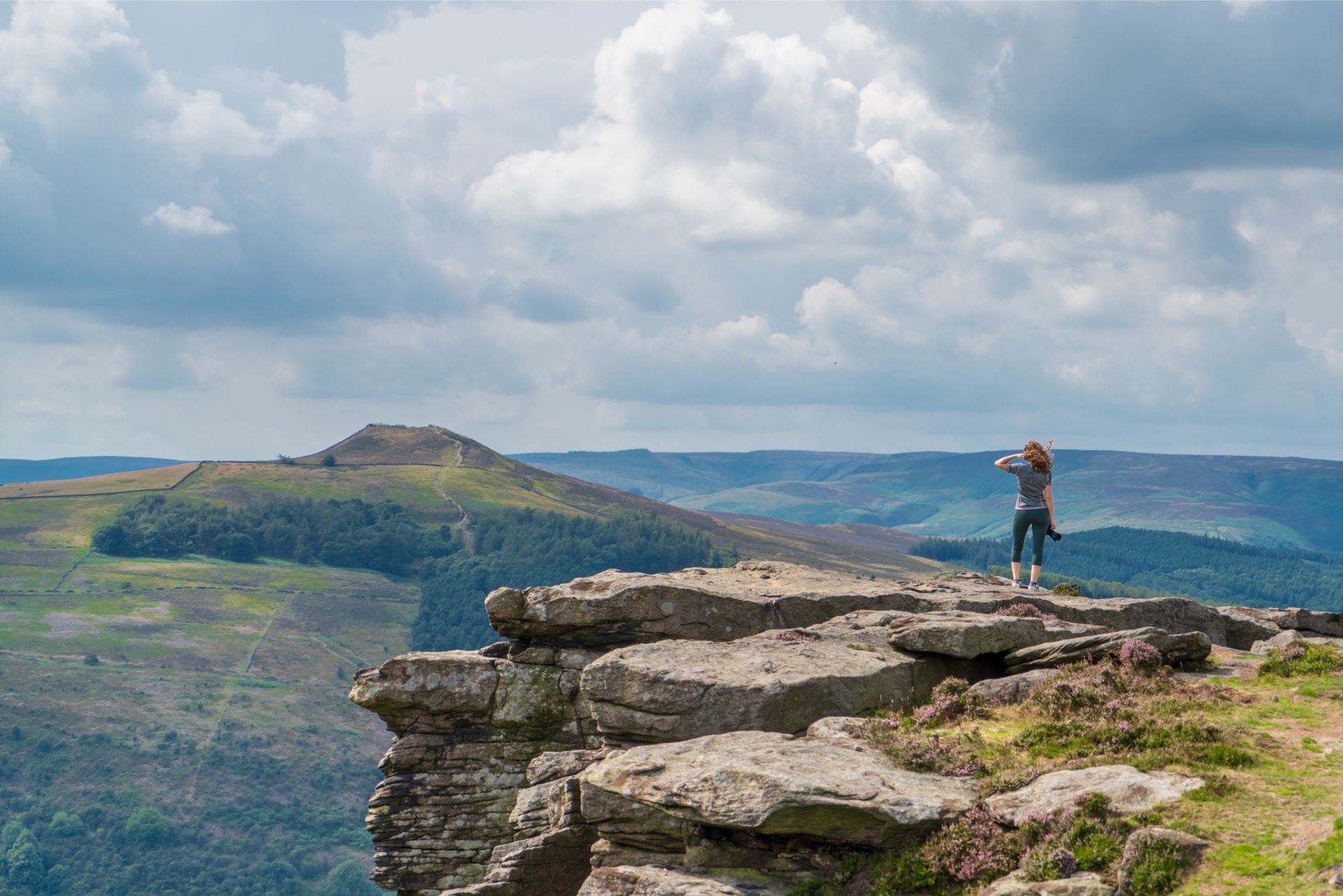 woman-standing-on-edge-of-cliff-looking-out-at-the-view-holding-a-camera-at-bamford-edge-in-the-peak-district