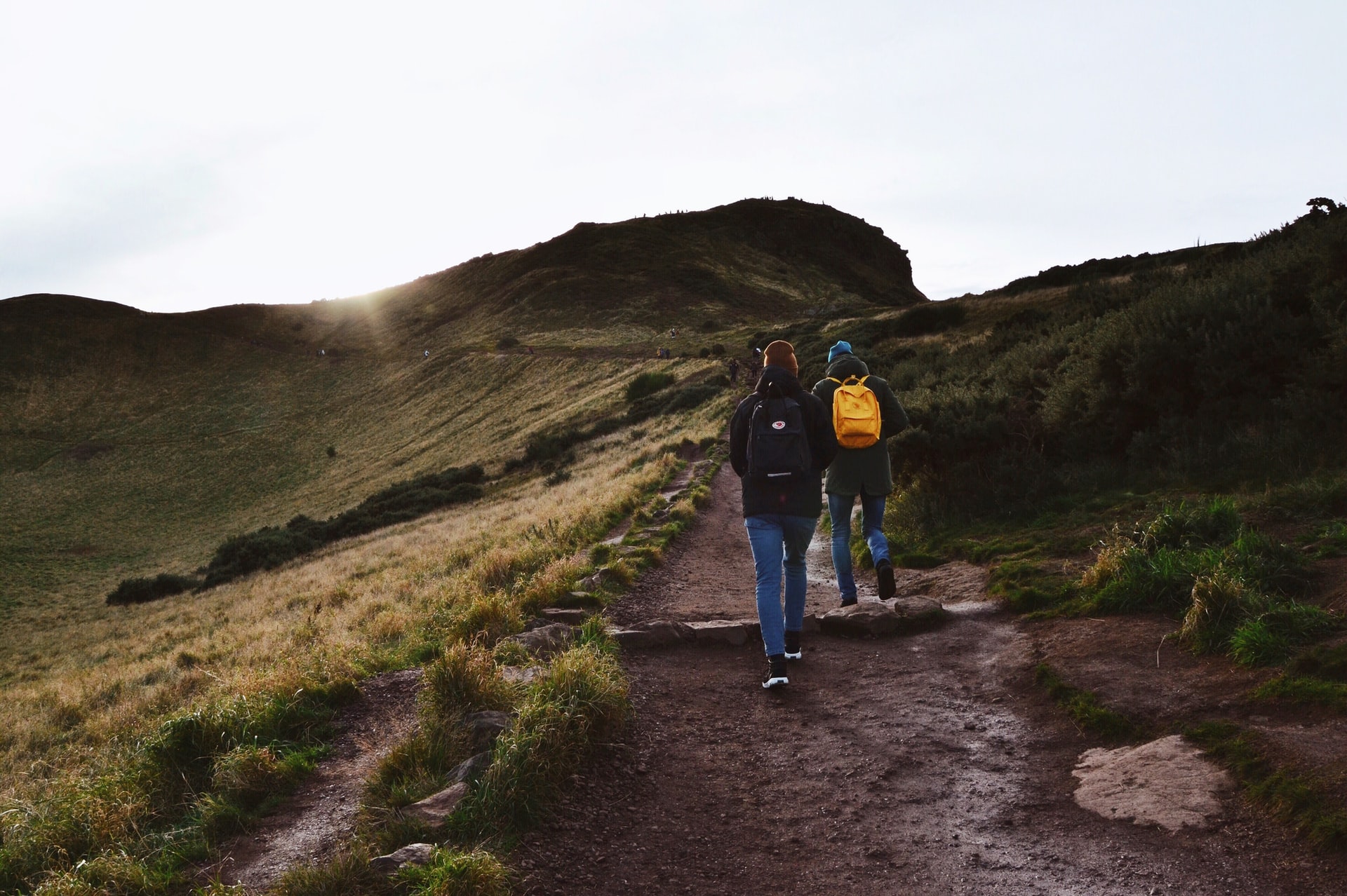two-people-wearing-jeans-a-beanie-and-backpack-climbing-up-a-hill-arthurs-seat-edinburgh