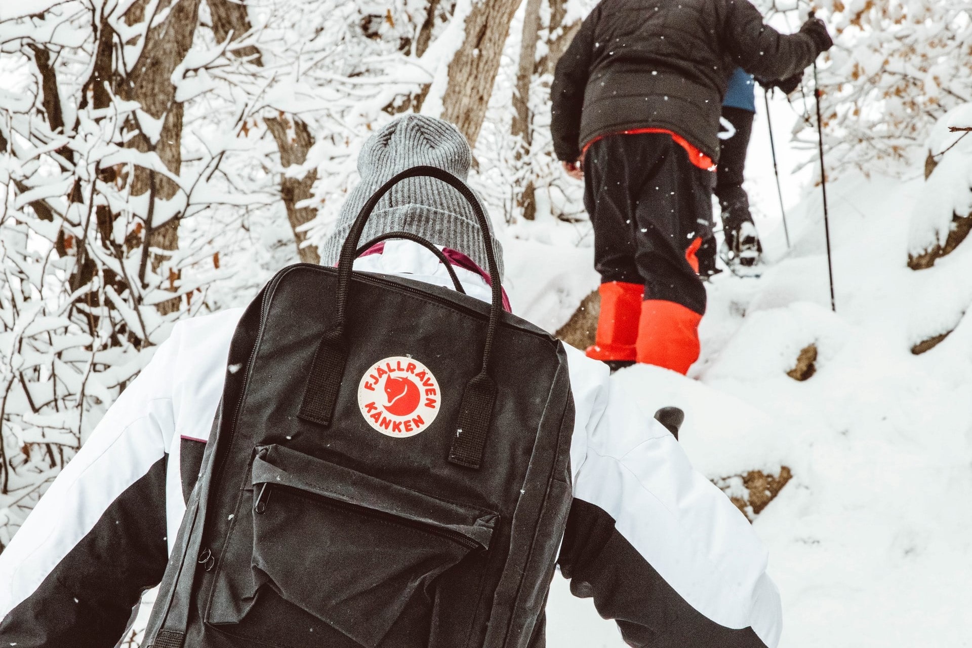 women-wearing-grey-beanie-and-fjallraven-kanken-backpack-climbing-mountain-with-walking-poles-in-snow-ski-season-packing-list