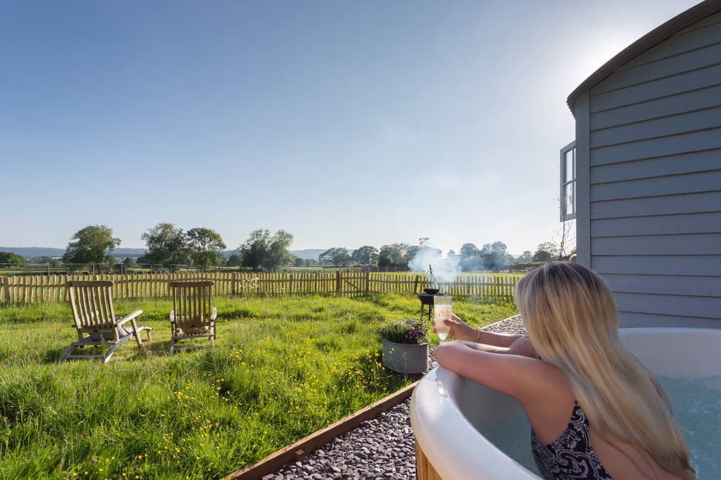 blonde-woman-in-hot-tub-with-glass-of-champagne-overlooking-field-by-grey-goose-shepherds-hut