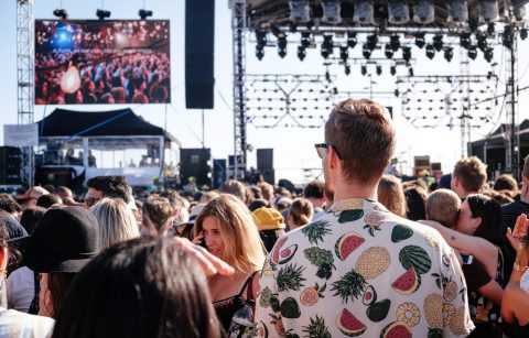 man-in-white-fruit-shirt-and-sunglasses-stood-in-festival-crowd-watching-a-band-in-daytime-packing-list-for-festival