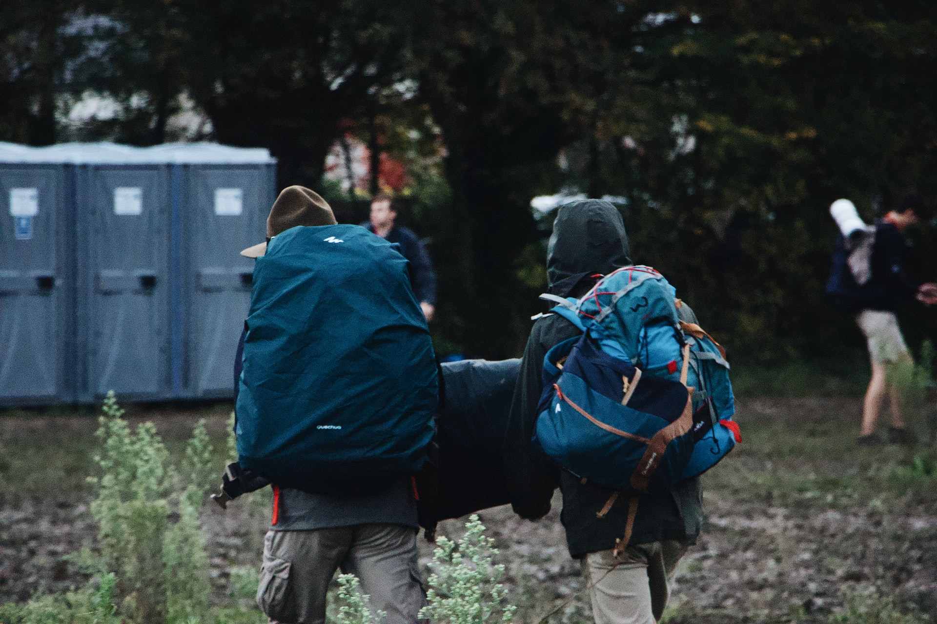 two-people-in-waterproofs-wearing-backpacks-walking-towards-portaloo-in-muddy-field-packing-list-for-festival