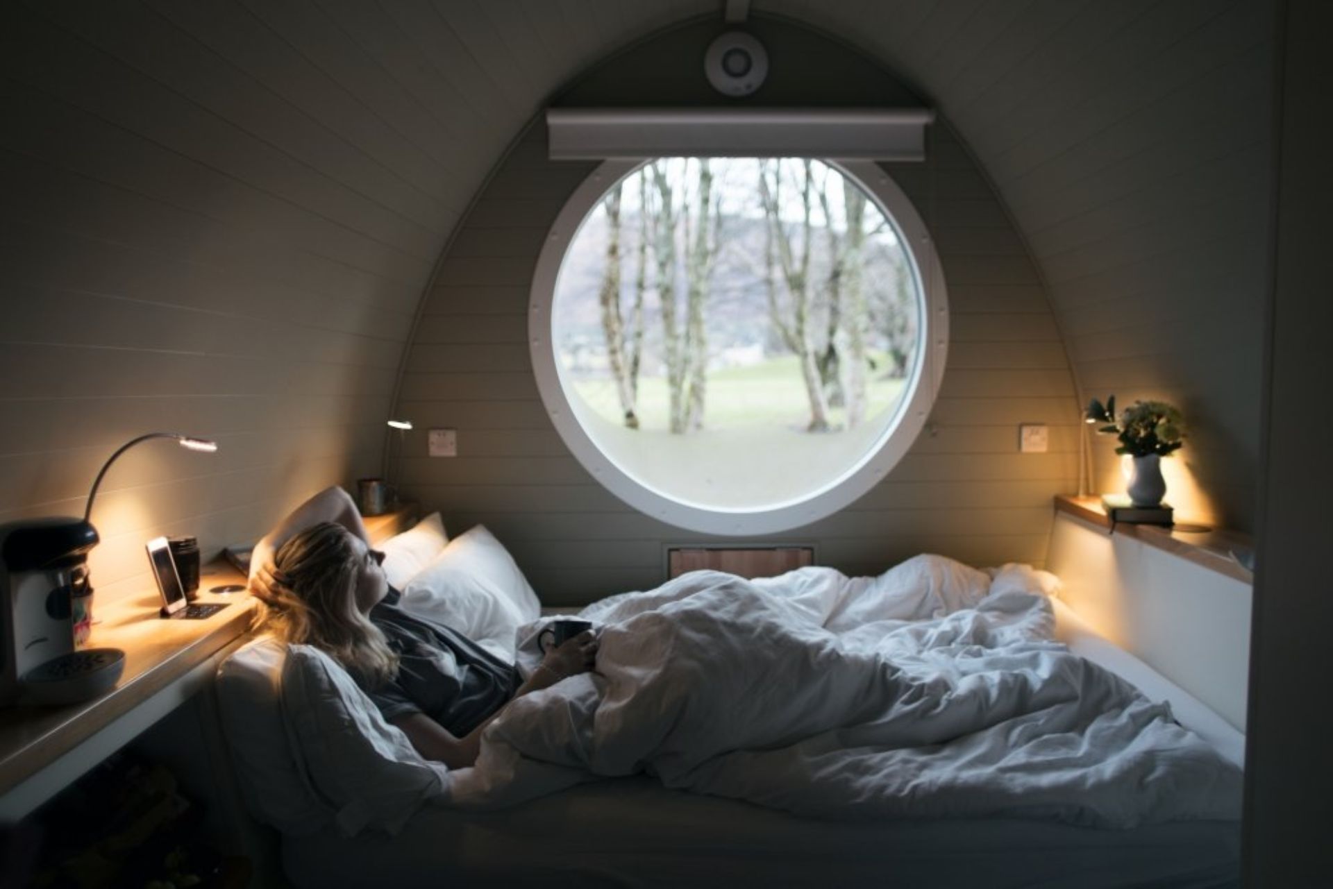 woman-lying-in-bed-in-riverbeds-lodge-glencoe-looking-out-of-a-round-window-lodges-with-hot-tubs-scotland