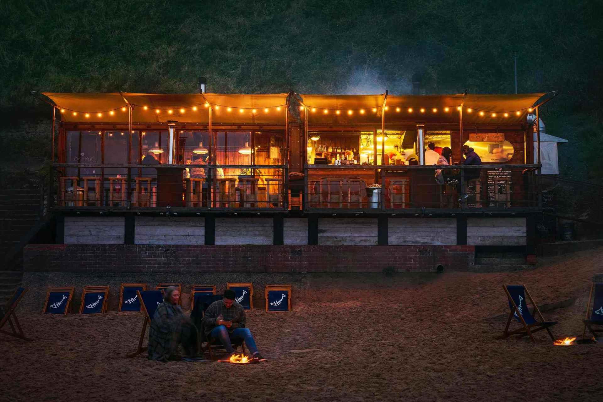 couple-sat-on-beach-by-fire-at-rileys-fish-shack-at-night