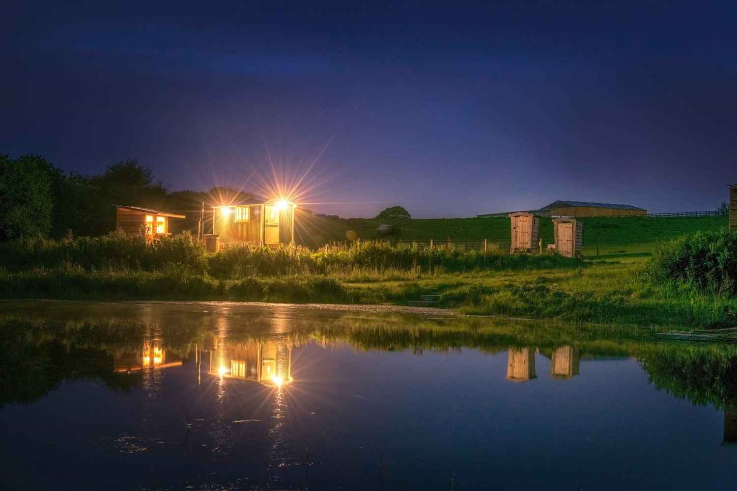 shepherds-hut-in-field-by-lake-lit-up-at-night-glamping-with-hot-tub-cornwall